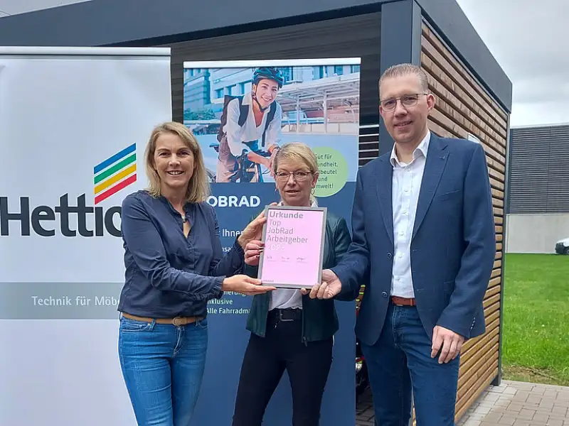 Monika Titze, von der Hettich Gruppe, steht zusammen mit einer Frau und einem Mann vor einem Poster mit dem Schriftzug Hettich und dem Hettich Logo. Sie alle tragen blaue Geschäftskleidung Monika Titze, von der Hettich Gruppe, steht zusammen mit einer Frau und einem Mann vor einem Poster mit dem Schriftzug Hettich und dem Hettich Logo. Sie alle tragen blaue Geschäftskleidung